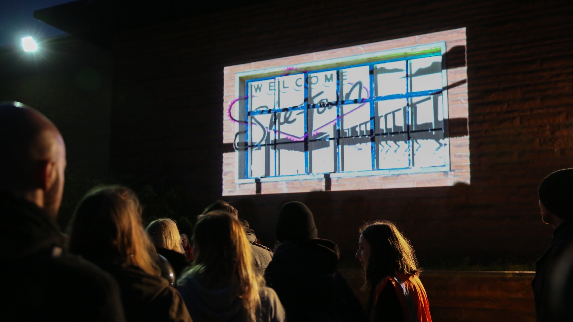 Photo of the event. The game is projected onto the large windows of the JCT factory, with the audience visible in the foreground.
