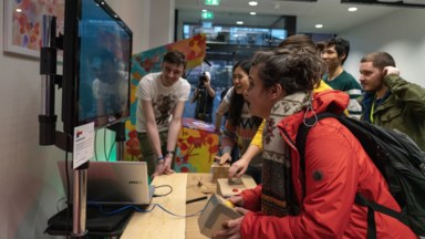 Photograph of someone playing the game Red Light Green Light, made for the jam. The game is a multiplayer game, with players alternately pressing buttons and hiding/revealing a light sensor to avoid detection. In the photo three players are watching the screen intently, themselves watched by a crowd of spectators.